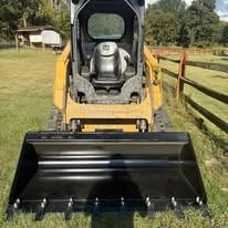 Yellow skid steer loader with front bucket attachment parked on grass field with wooden fence in background