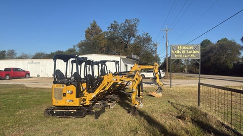 Yellow compact excavator parked on grass near a commercial lot